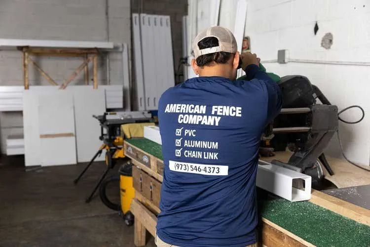 Man in blue shirt from American Fence Company uses a saw to cut a white fence post inside a workshop.