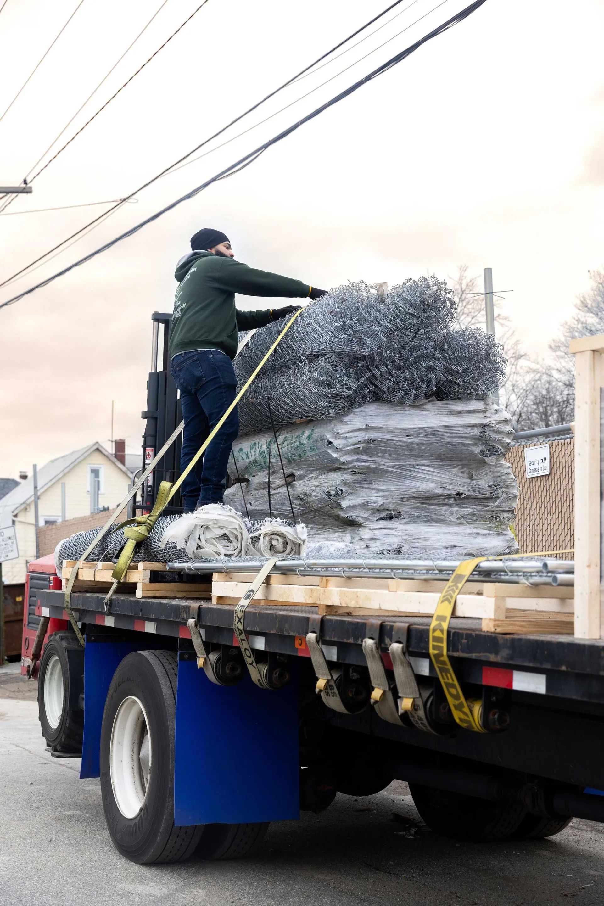 A worker secures a load of metal fencing on a flatbed truck; outdoor setting, overcast sky.