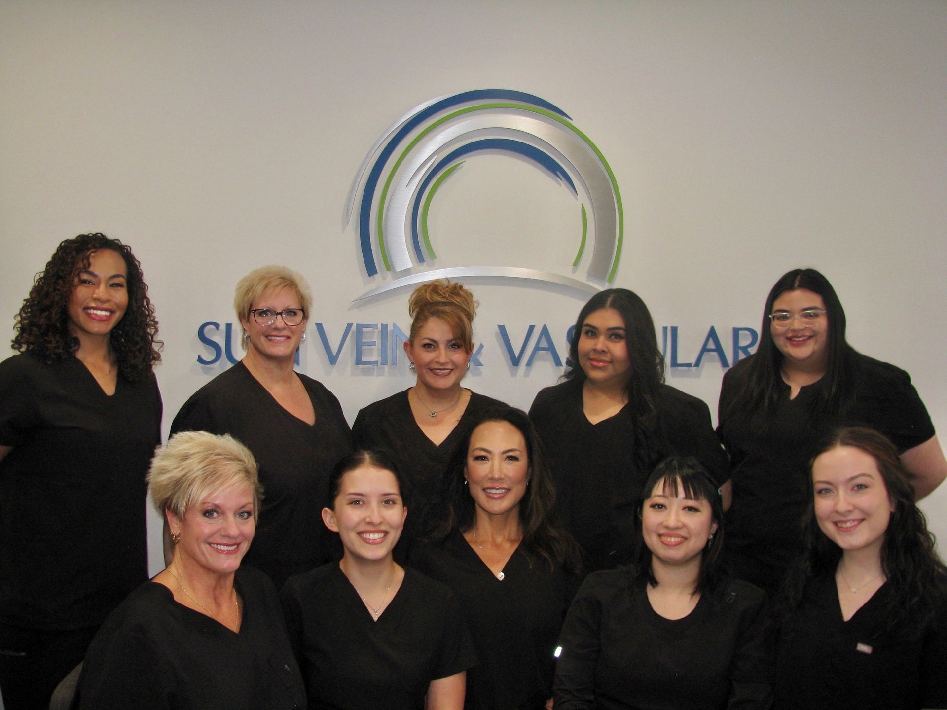 Staff of a medical office smiling together, wearing black tops in front of a logo.