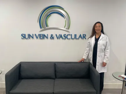 Woman in lab coat stands by a gray couch, below a "Sun Vein & Vascular" sign on a white wall.