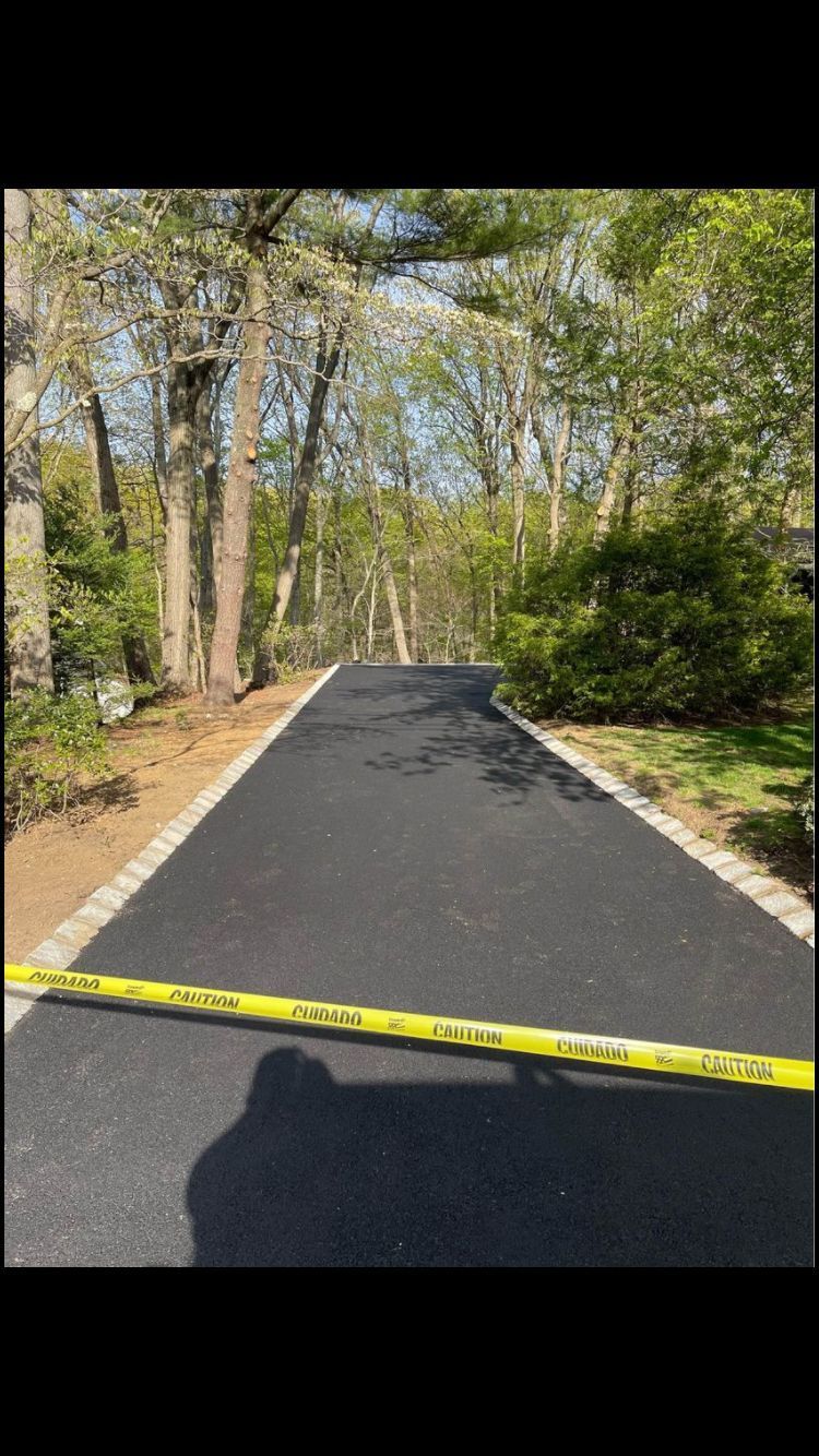 Newly paved asphalt driveway with yellow caution tape, bordered by stone and flanked by trees.