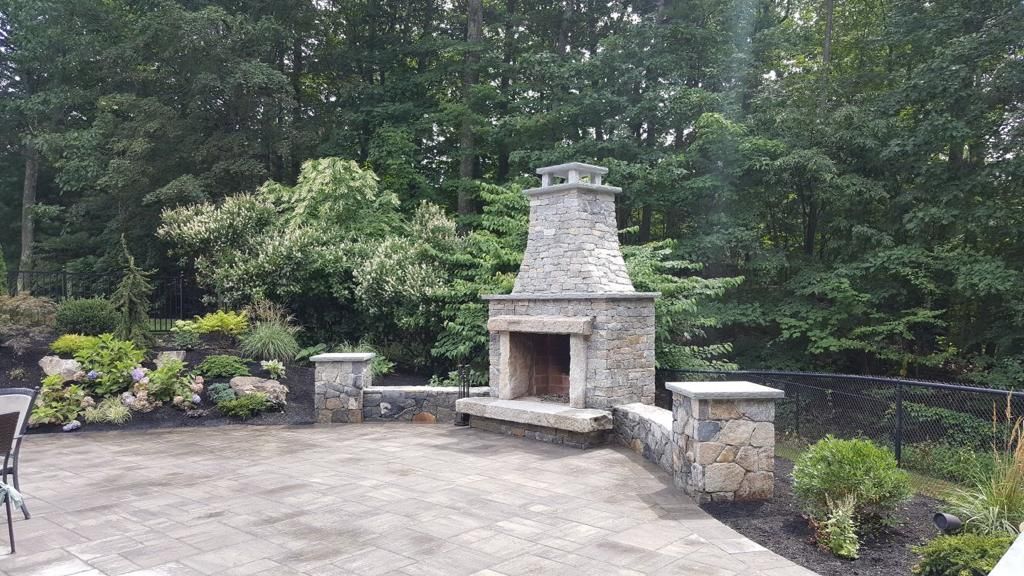 A stone fireplace is in the middle of a patio surrounded by trees.