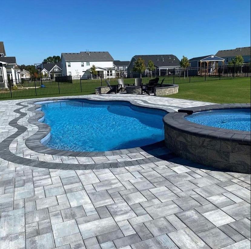 Swimming pool and spa with blue water, surrounded by gray pavers. Houses and lawn in background.