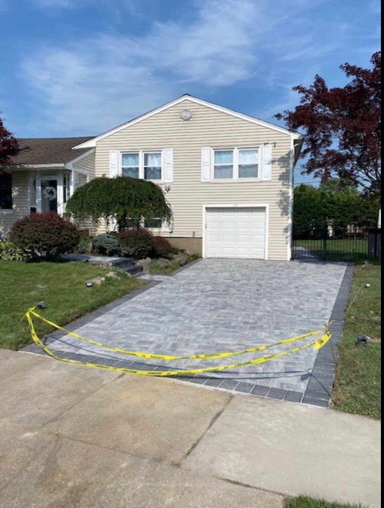 House with new gray paver driveway, edged with dark pavers, bordered by grass and caution tape.