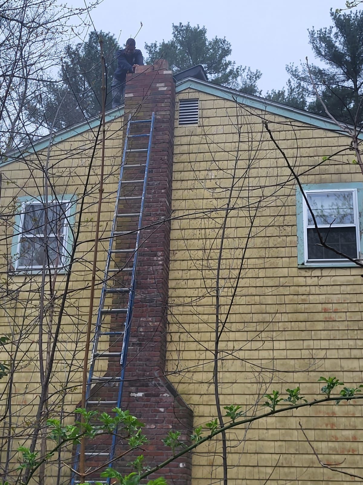 A man on a ladder is working on a chimney on the side of a house.