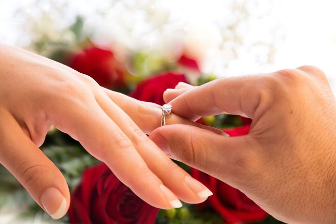 Hand placing a diamond ring on another hand, red roses in the background.