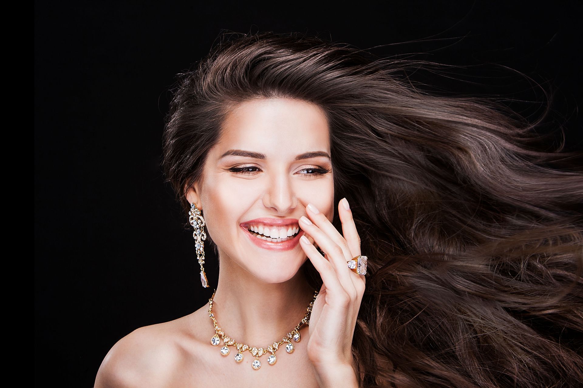 Woman with long brown hair, wearing jewelry, smiling, against a black background.