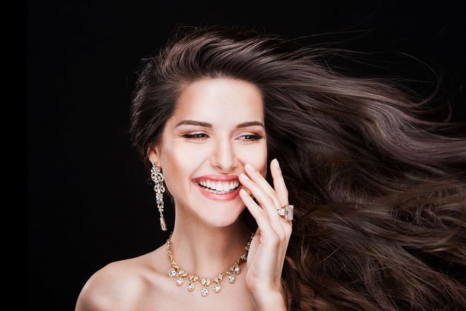 Woman with long brown hair, wearing jewelry, smiling, against a black background.