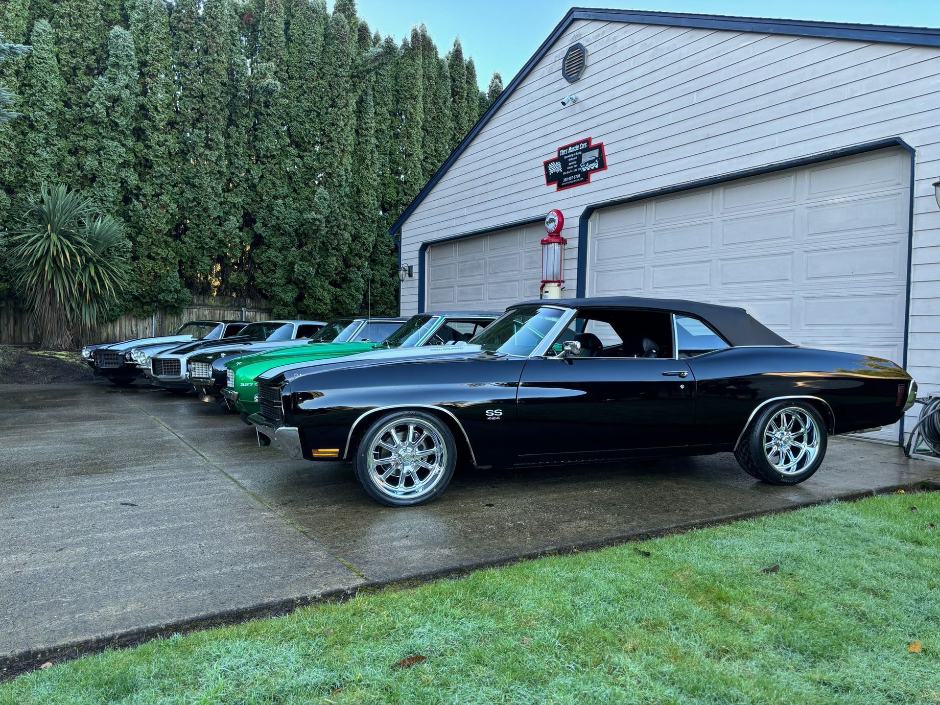 Five classic cars parked in front of a garage; black convertible in front, others silver and green.