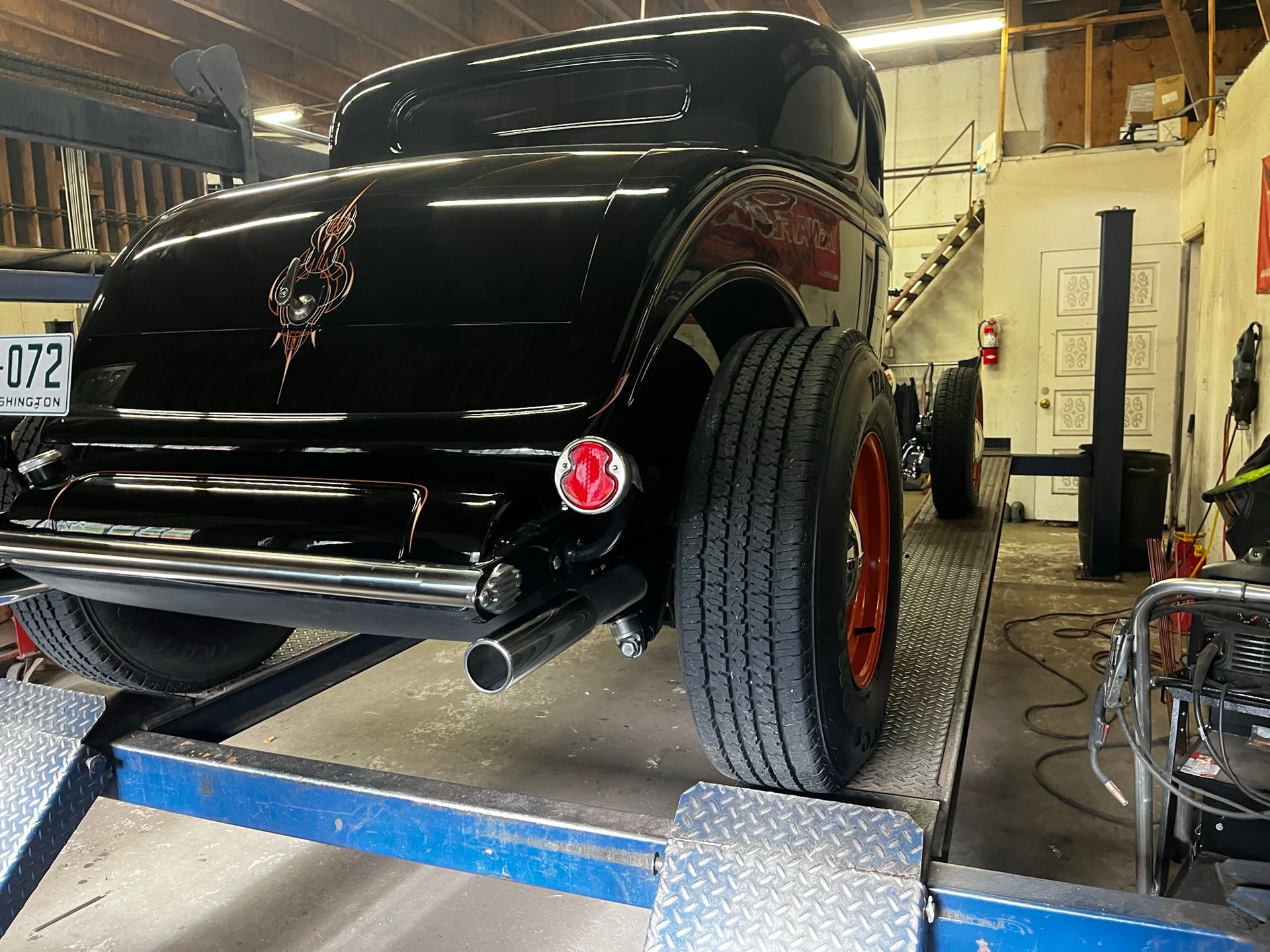 Black hot rod car on a lift in a garage, with red wheels and exhaust pipes.