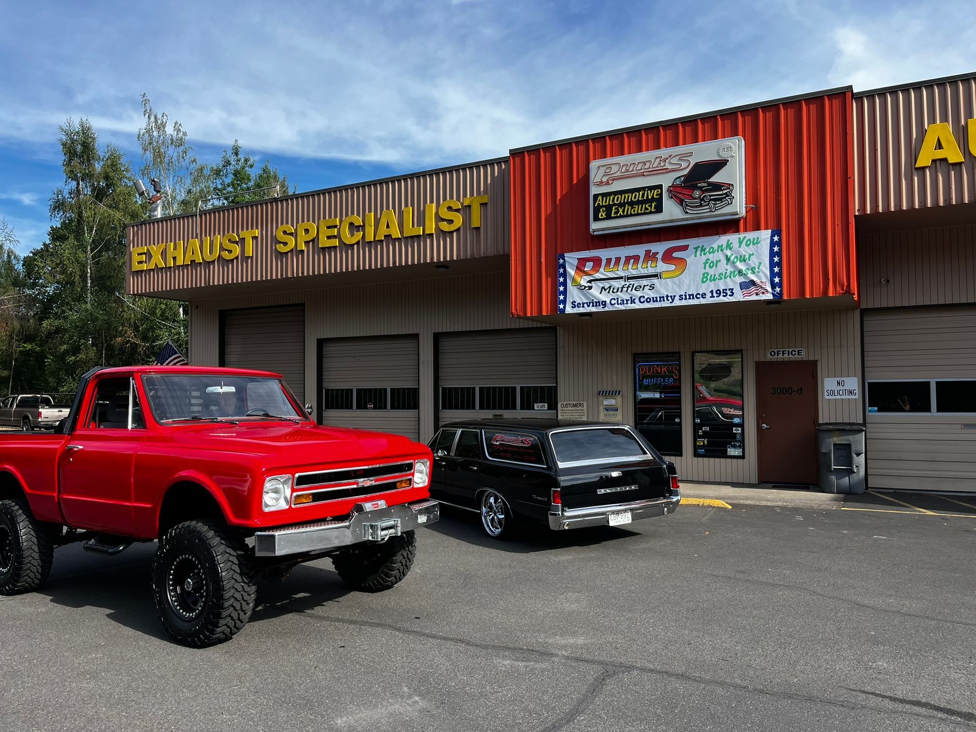 Red truck and black car parked in front of an exhaust specialist shop.