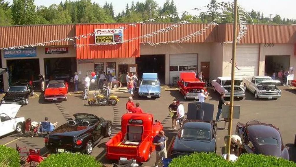 Classic car show outside auto shop. Red, blue, and black vehicles on display. People gather around.