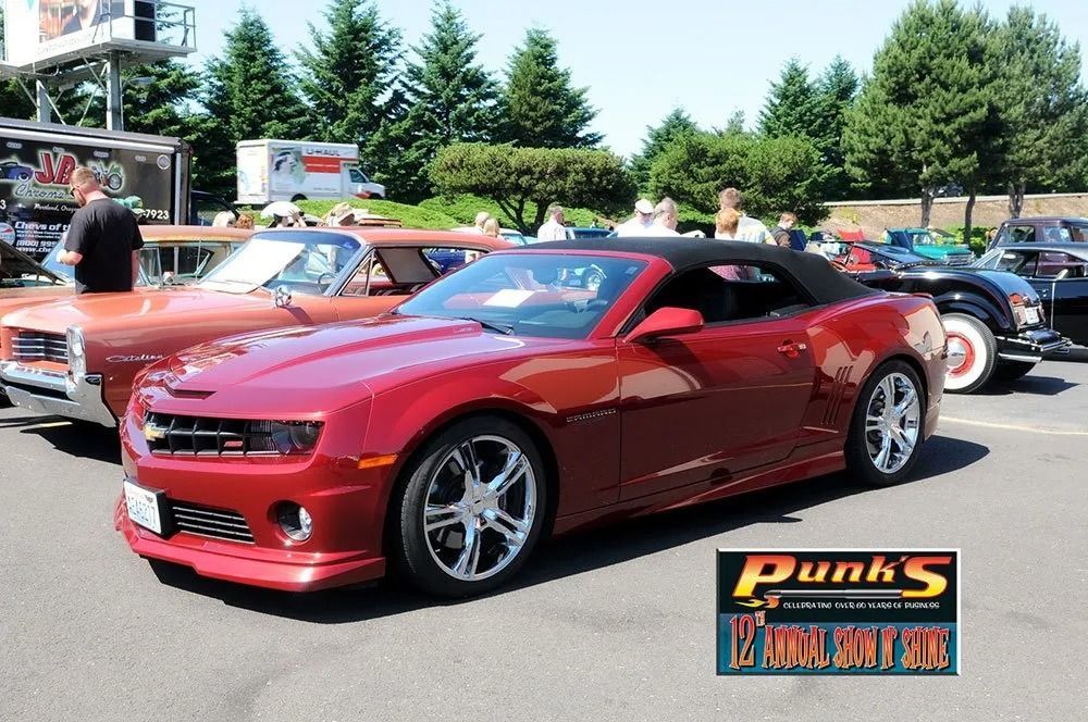 Red convertible Camaro at a car show, sunny day. Other classic cars and people in the background.