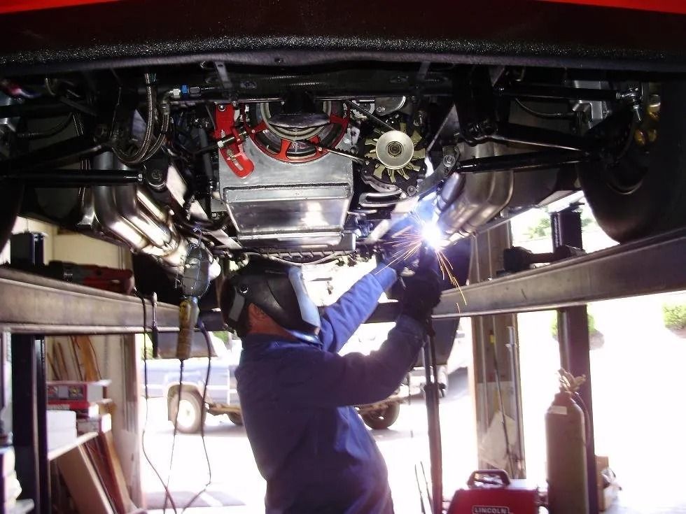 Mechanic welding underneath a red car raised on a lift; welding sparks visible, garage setting.