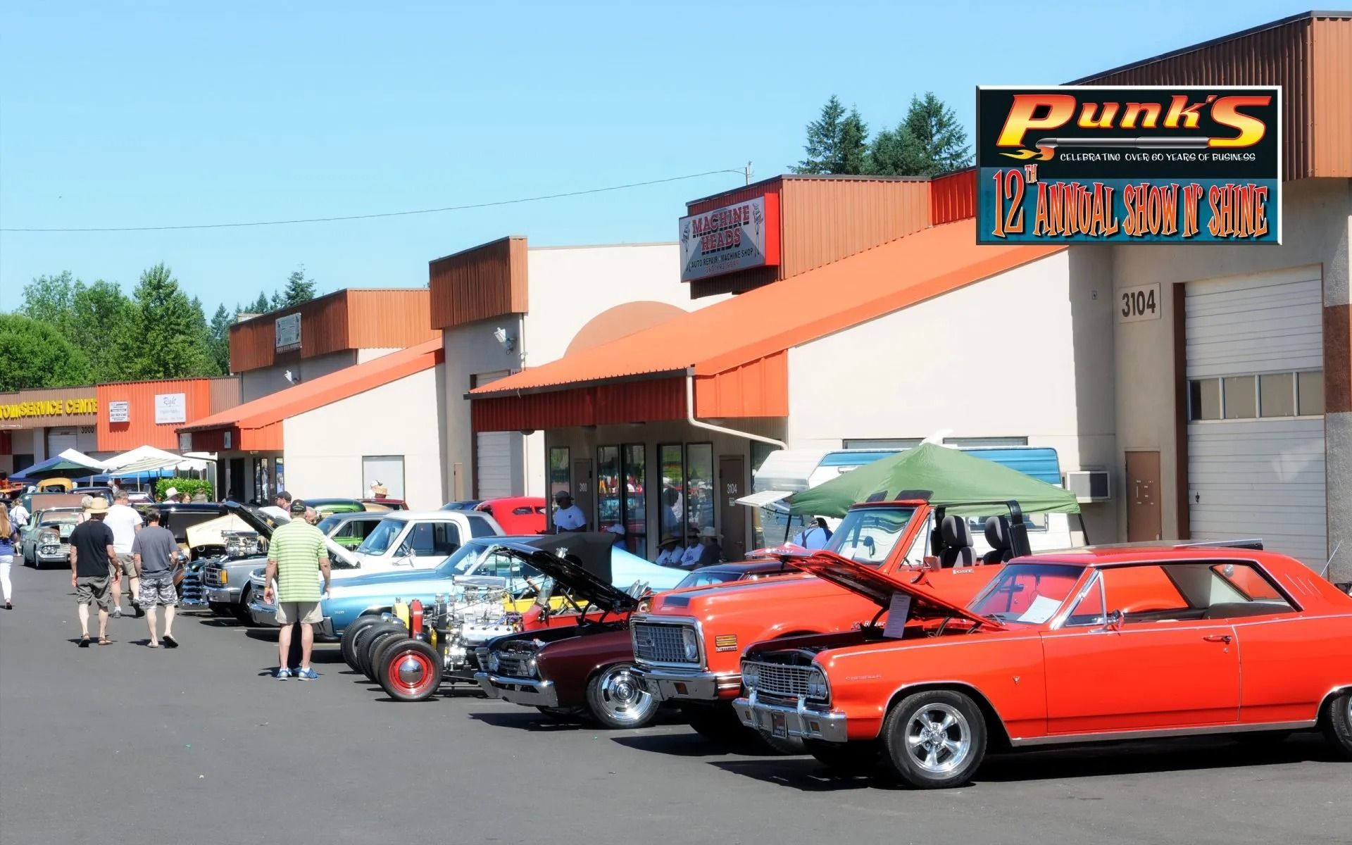 Classic cars on display at Punk's Hot Rods. People are walking around and looking at the vehicles in front of a building.