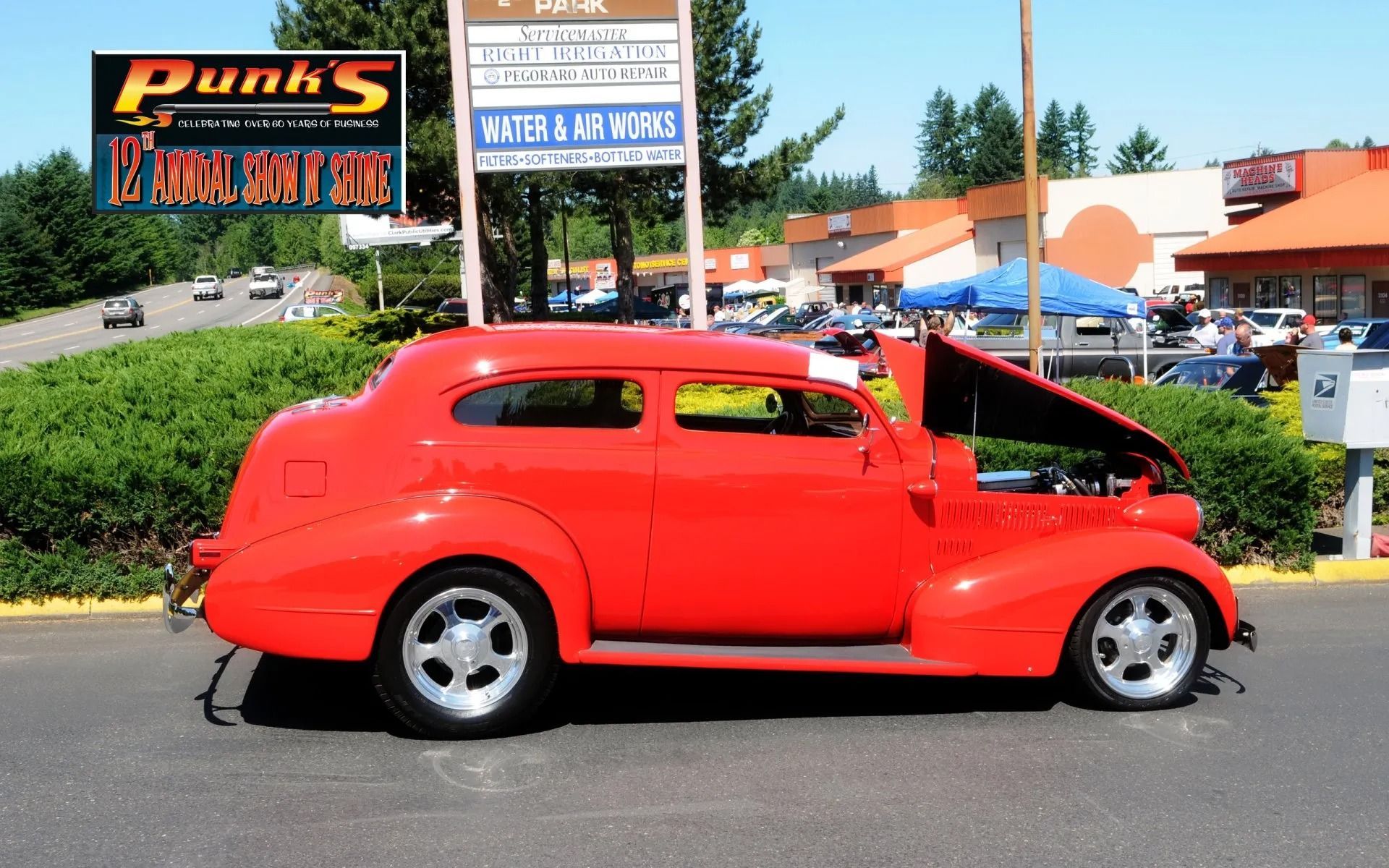 Red vintage car with hood up at an outdoor car show, in front of shops.