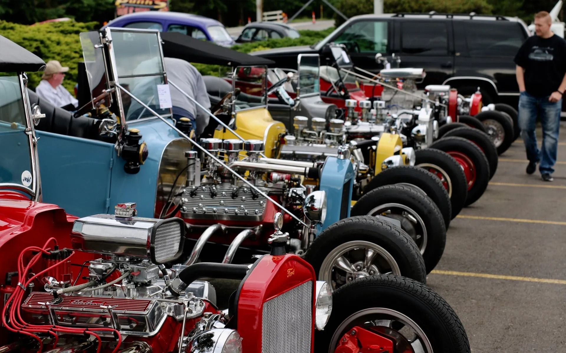 Line of vintage, customized hot rods at an outdoor car show; engines on display, man walks by.