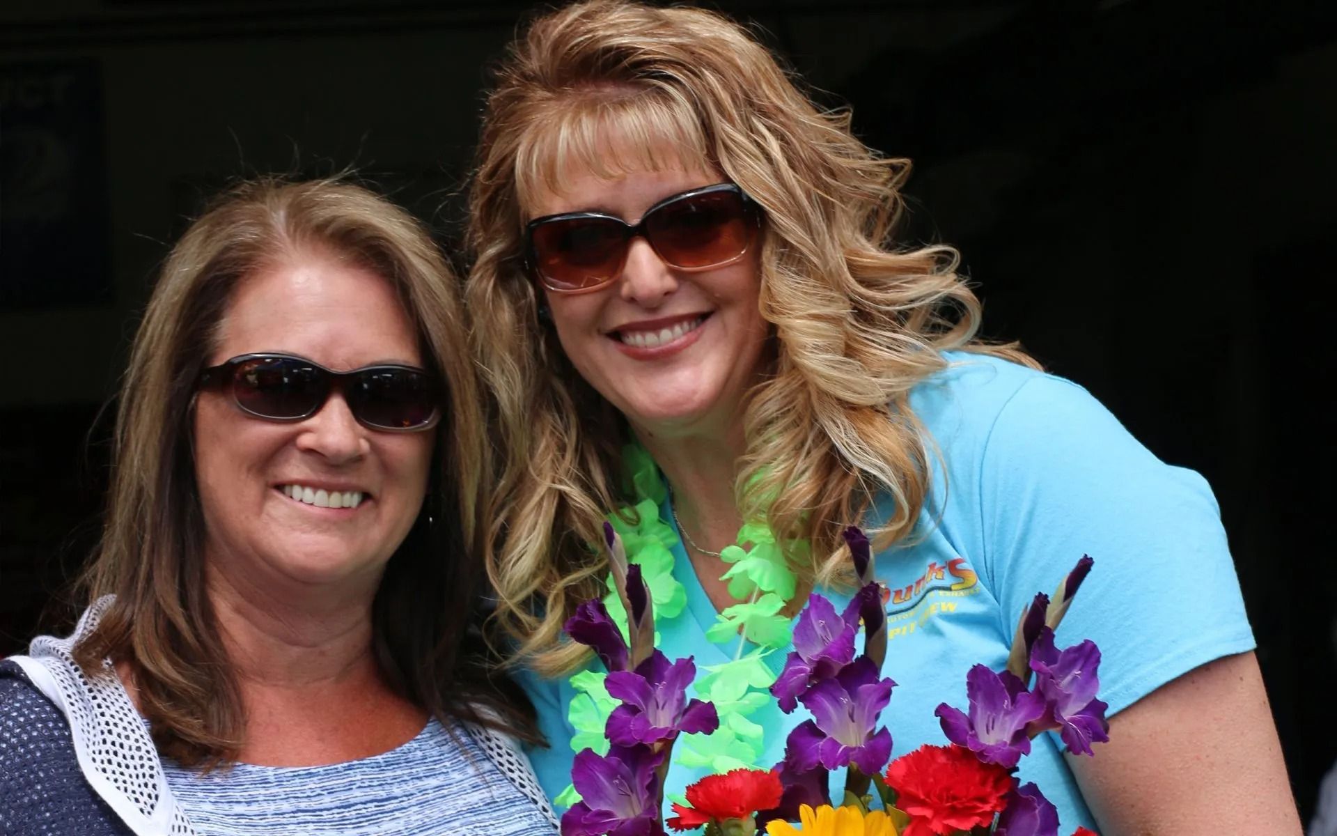 Two women smiling, one wearing sunglasses, a lei, and holding flowers.