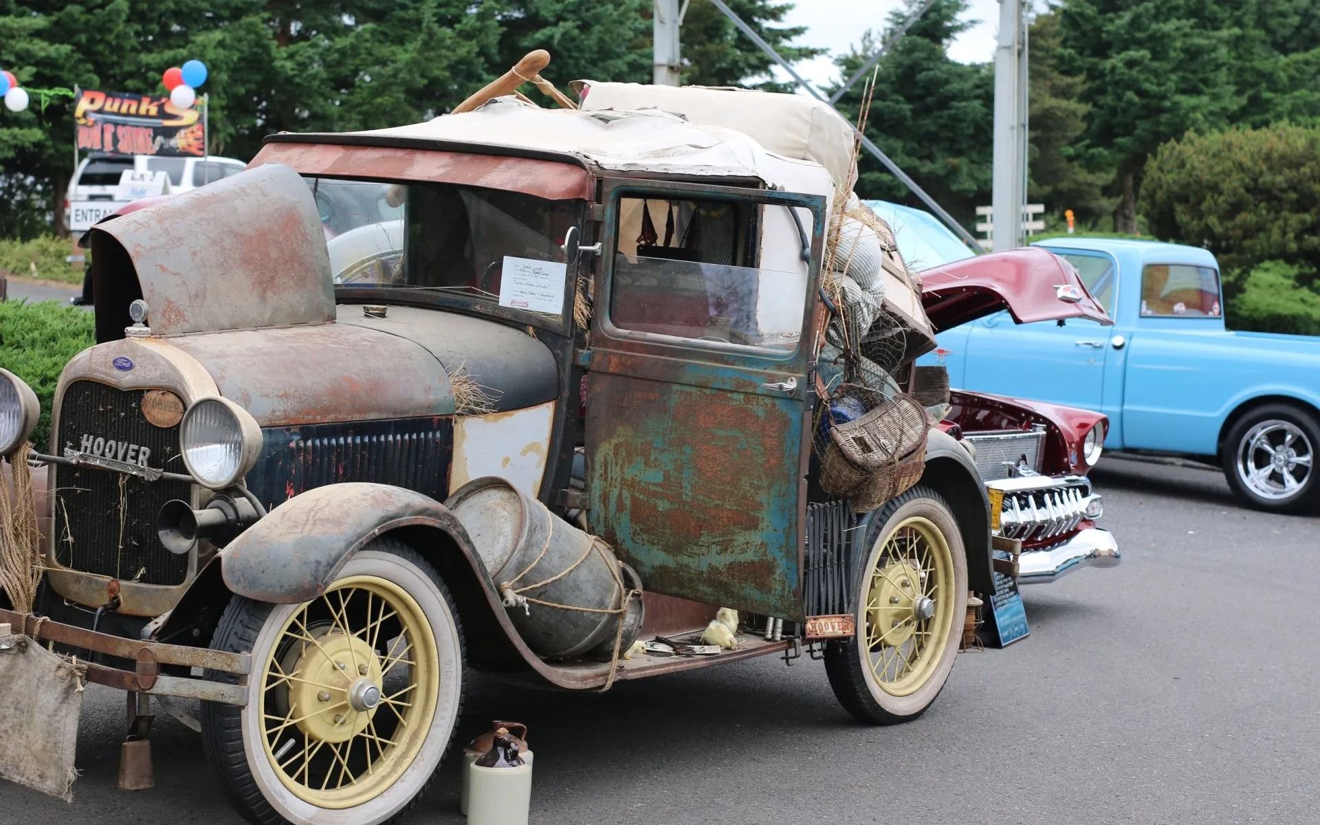 Rusty vintage car with yellow wheels parked, blue truck in background.