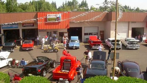 Classic cars on display outside an auto repair shop. People milling around, sunny day.