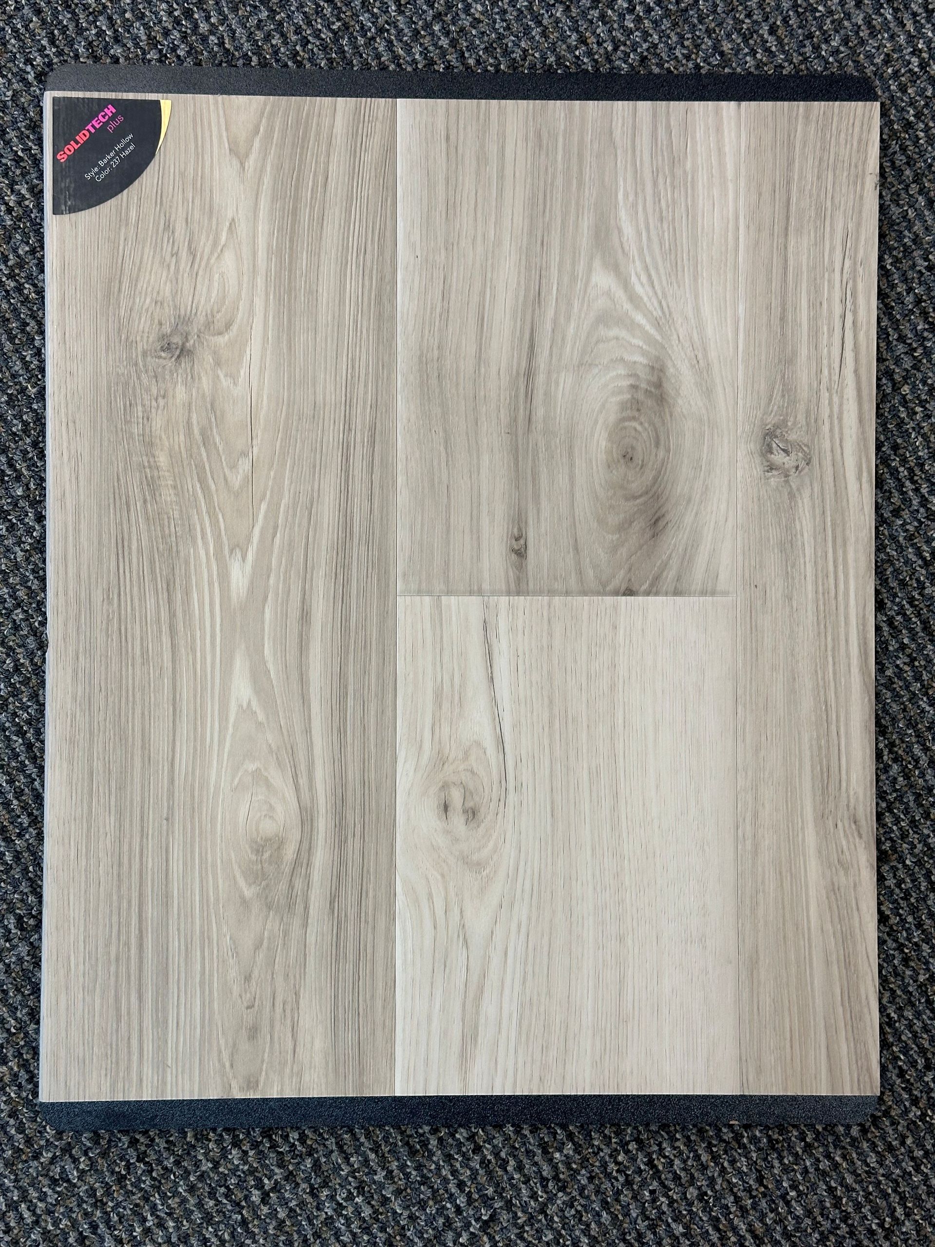 Light-brown wood flooring sample, close-up view. Displayed on a dark gray carpeted surface.