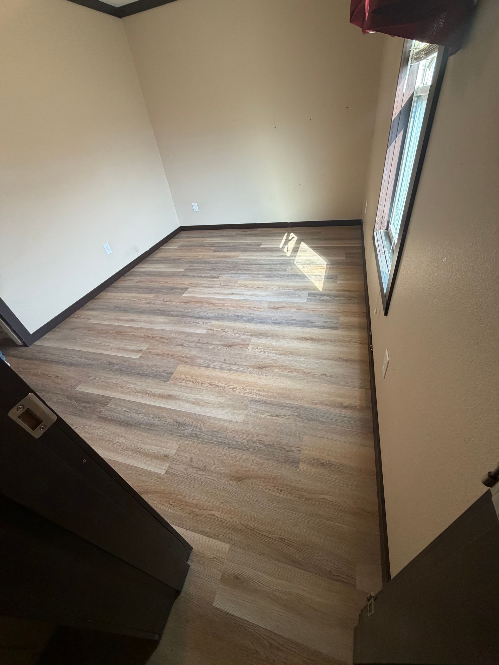Empty room with wood-look flooring and a window. Beige walls, dark trim, and a ray of sunlight.