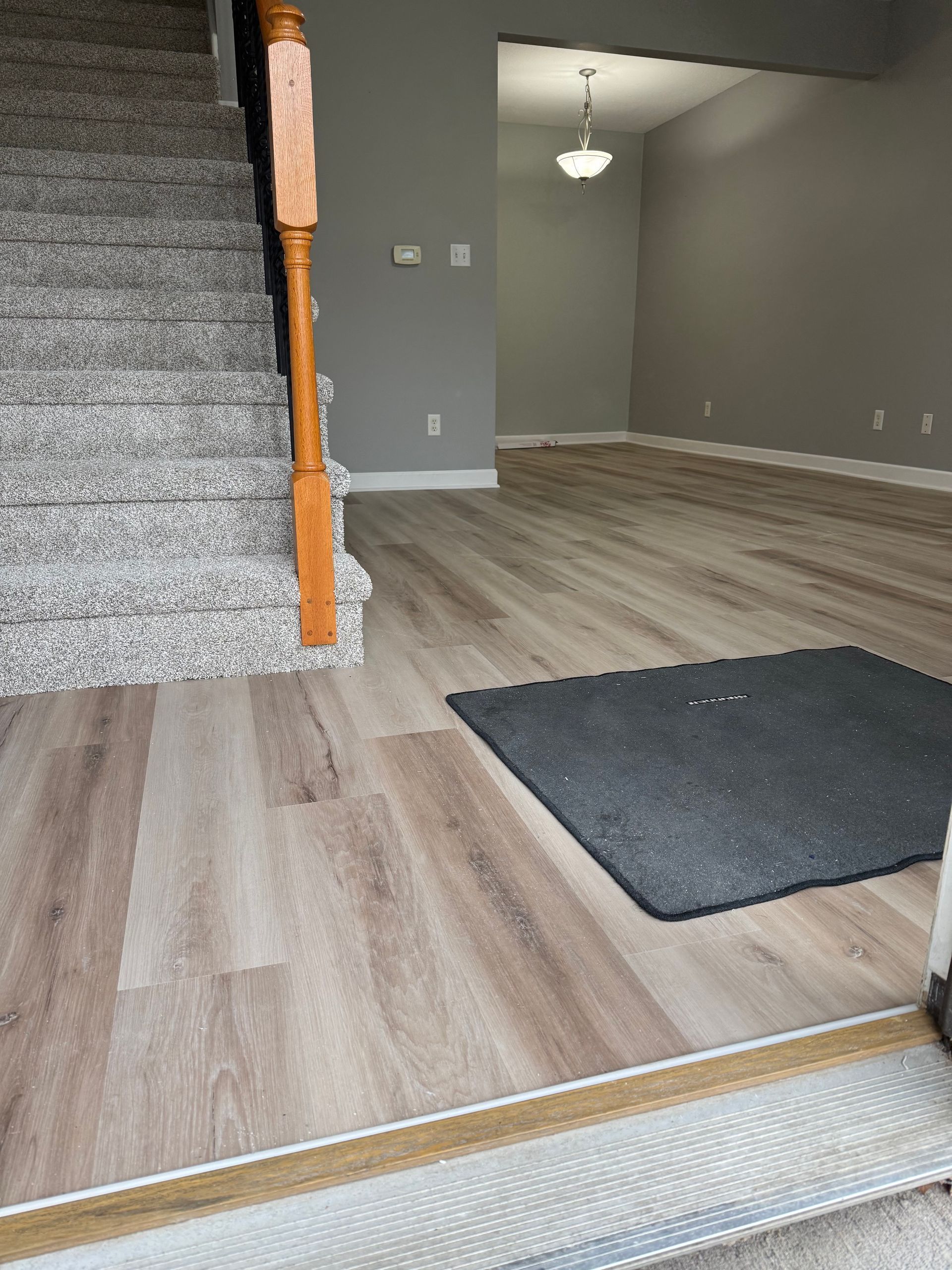 Staircase with carpet steps, wood railing, next to wood-look flooring. Gray walls and a doorway into another room. Black floor mat.