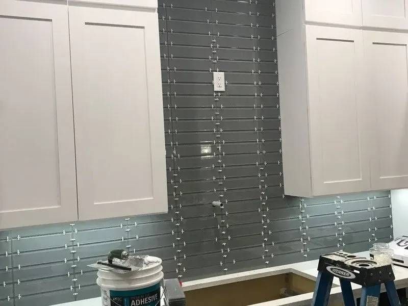 Kitchen backsplash under construction with gray tiles, white cabinets, and a partially installed tile installation.