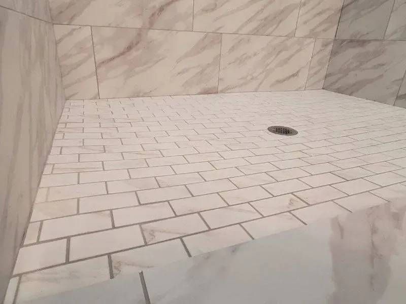 White tiled shower interior with a drain in the center. The walls and floor are covered in rectangular tiles, with a marble pattern on the walls.