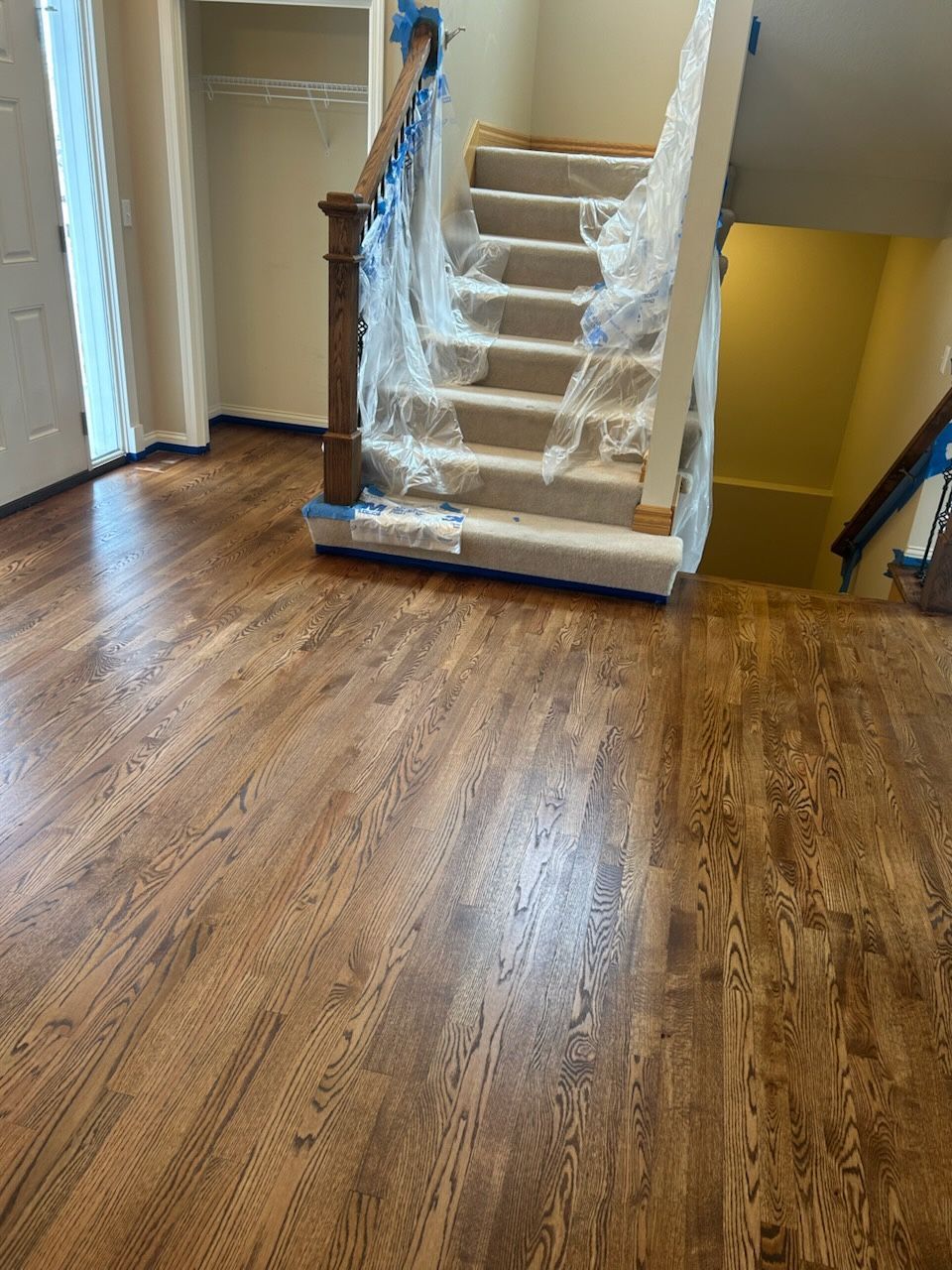 Hardwood floor in a home with a staircase covered in plastic. Staircase has a brown banister.