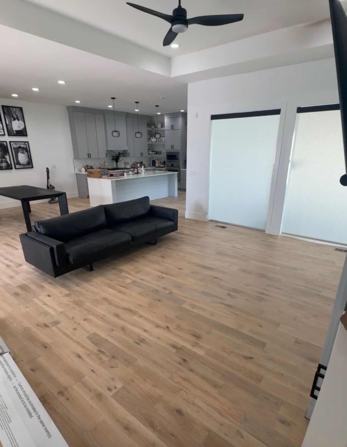 Open-concept living space with light wood floors, a black sofa, and a kitchen in the background. 