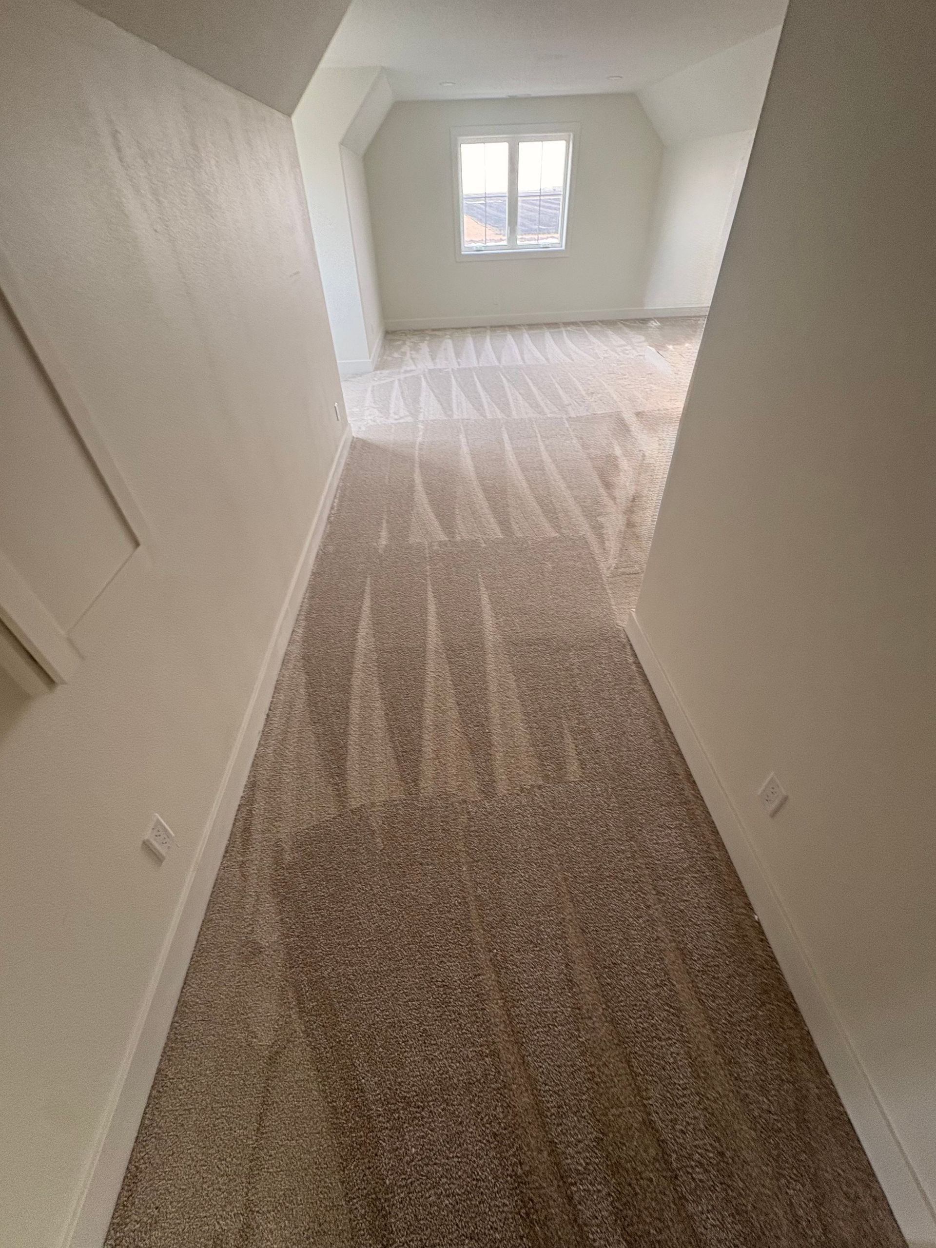 A hallway with beige carpet leads to a room with a window, white walls, and a sloped ceiling.