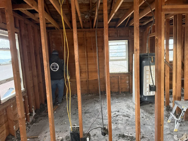 A person stands in a construction zone inside a wood-framed house under renovation with exposed wiring and open walls.