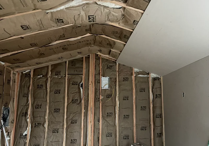 Interior view of a room under construction, showing wood wall studs, fiberglass insulation, and partially installed drywall.