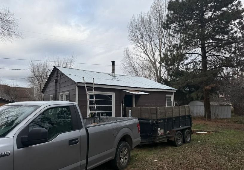 A brown house with a new metal roof under construction, with a silver pickup truck and utility trailer parked nearby.