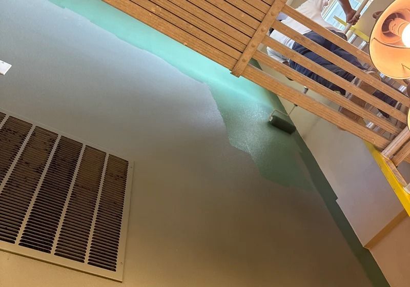 A low-angle view shows a person using a roller to paint a wall with light green paint next to a wood-slatted ceiling.