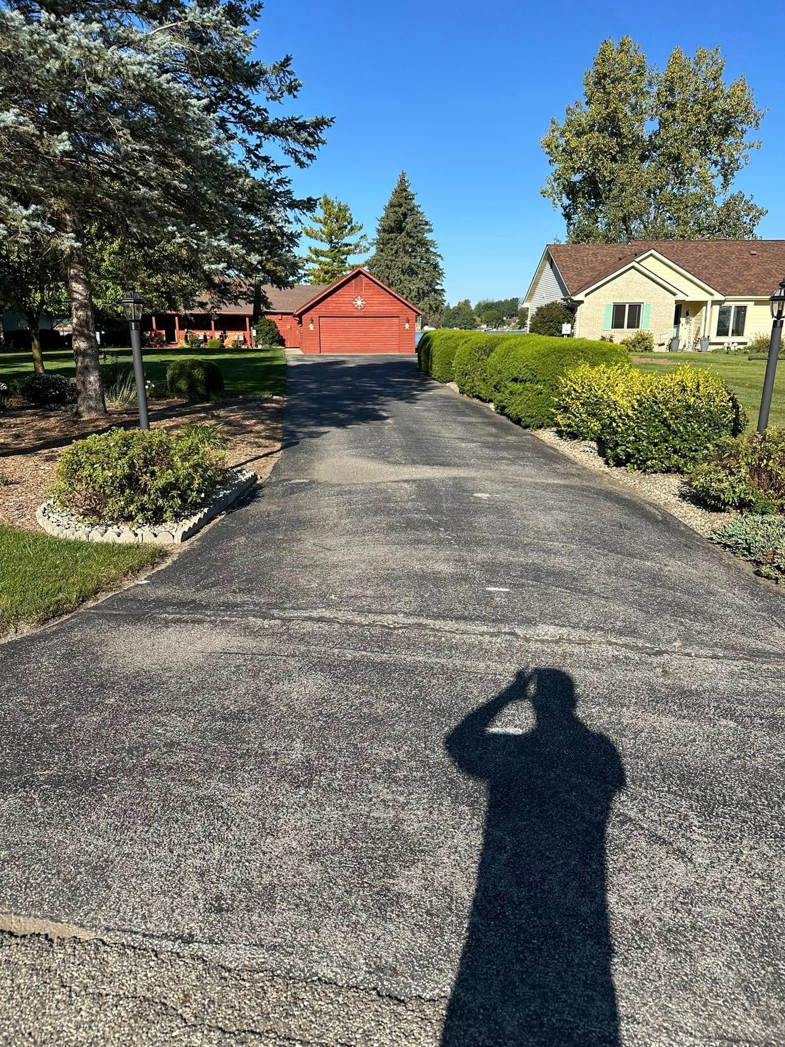 Asphalt driveway leads to a red garage and houses under a clear sky. Shadow of person taking photo visible.