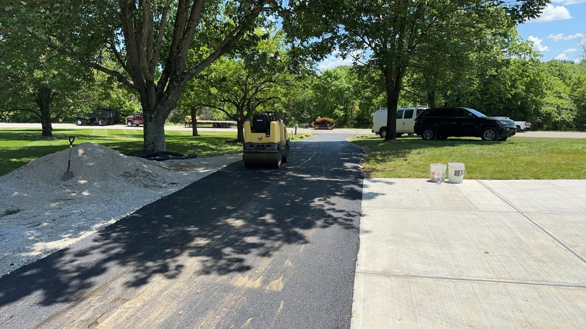 Asphalt paving a driveway; roller machine in use; trees and a car in the background.