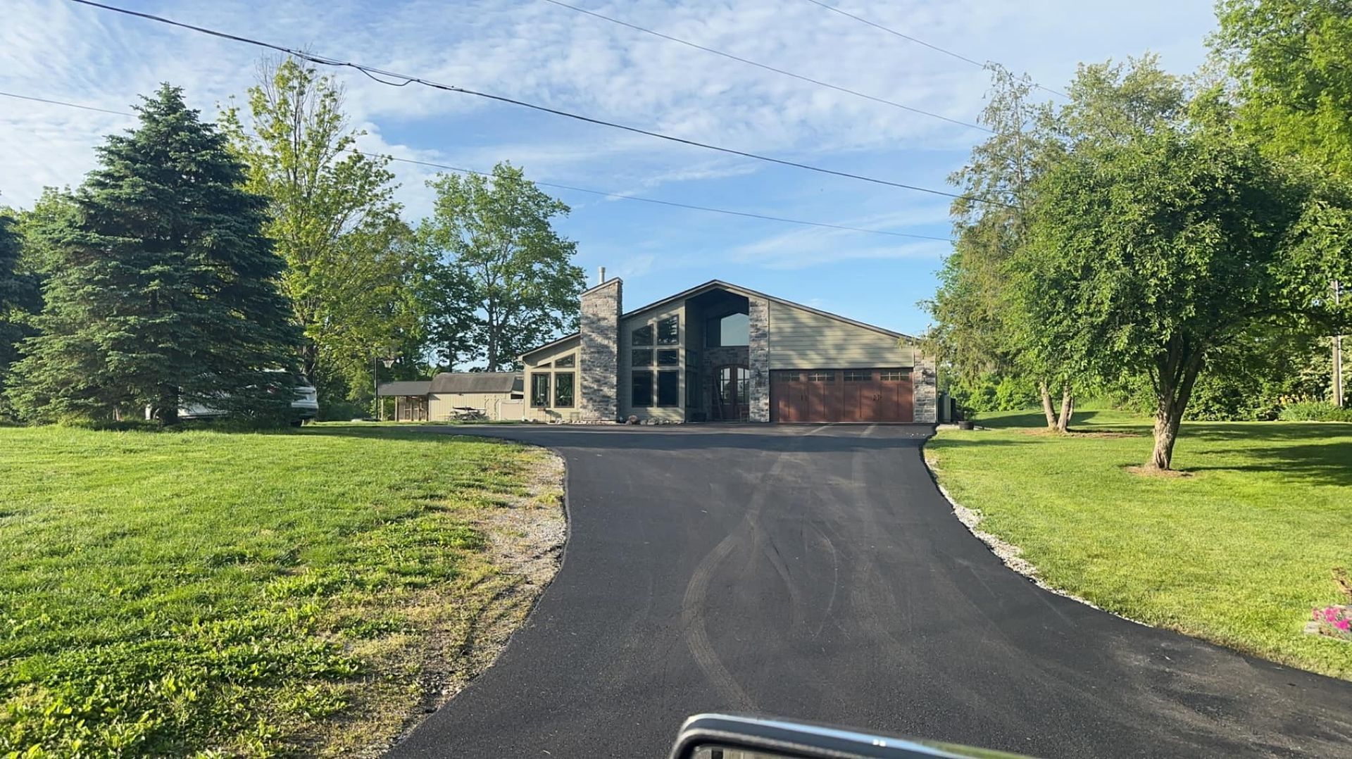 A modern, one-story house with a long paved driveway under a blue sky. Green trees and grass surround.