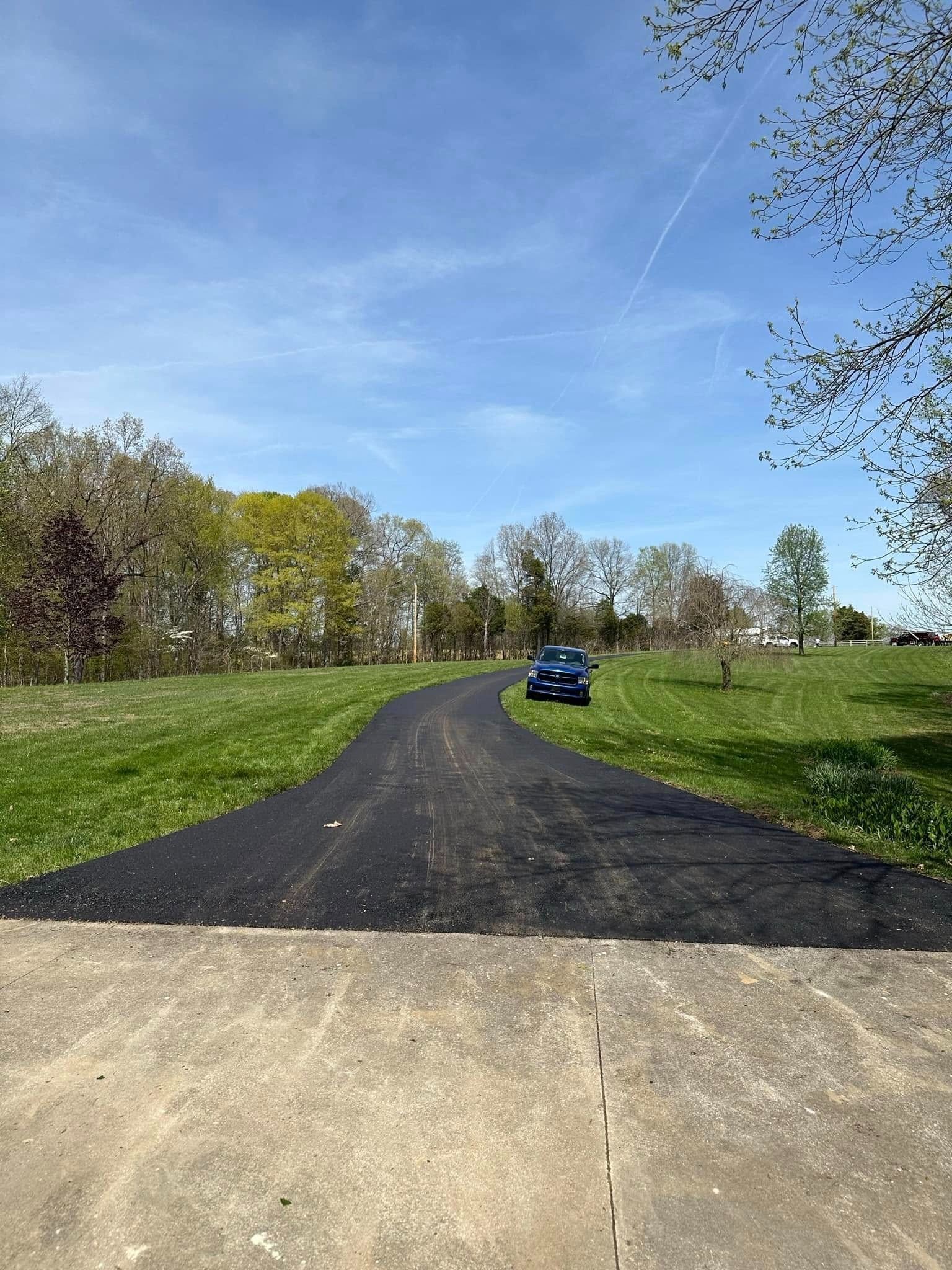Asphalt driveway leading past a parked blue car through a grassy field under a blue sky.