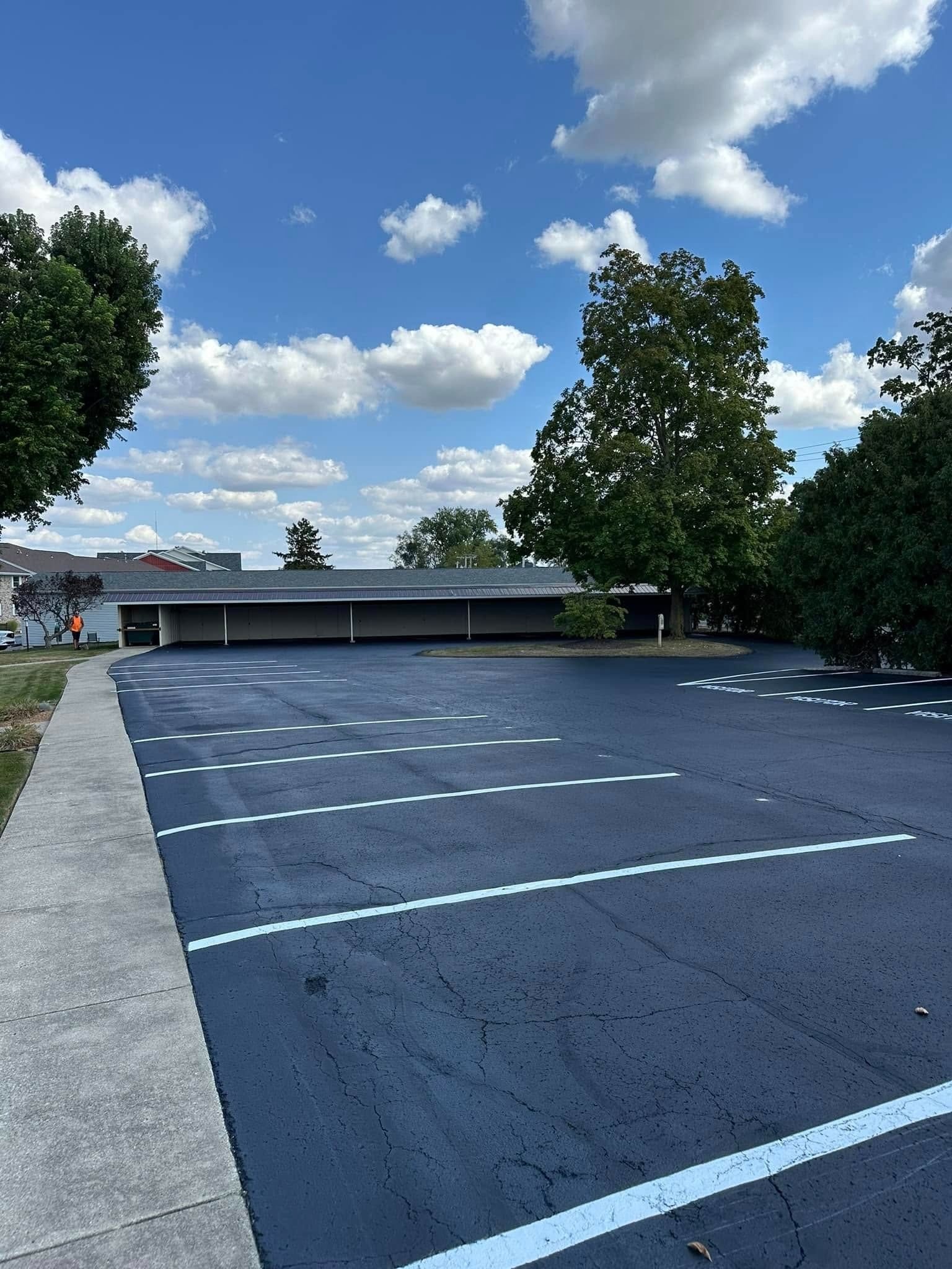 Newly paved black parking lot with white painted parking space lines. Trees, buildings, and blue sky with clouds in background.