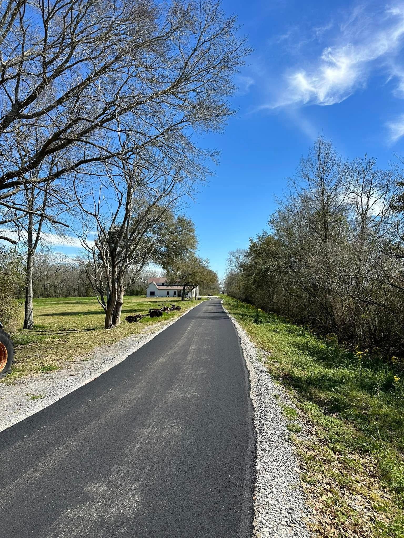 Asphalt path through a grassy area, trees lining sides, blue sky overhead.