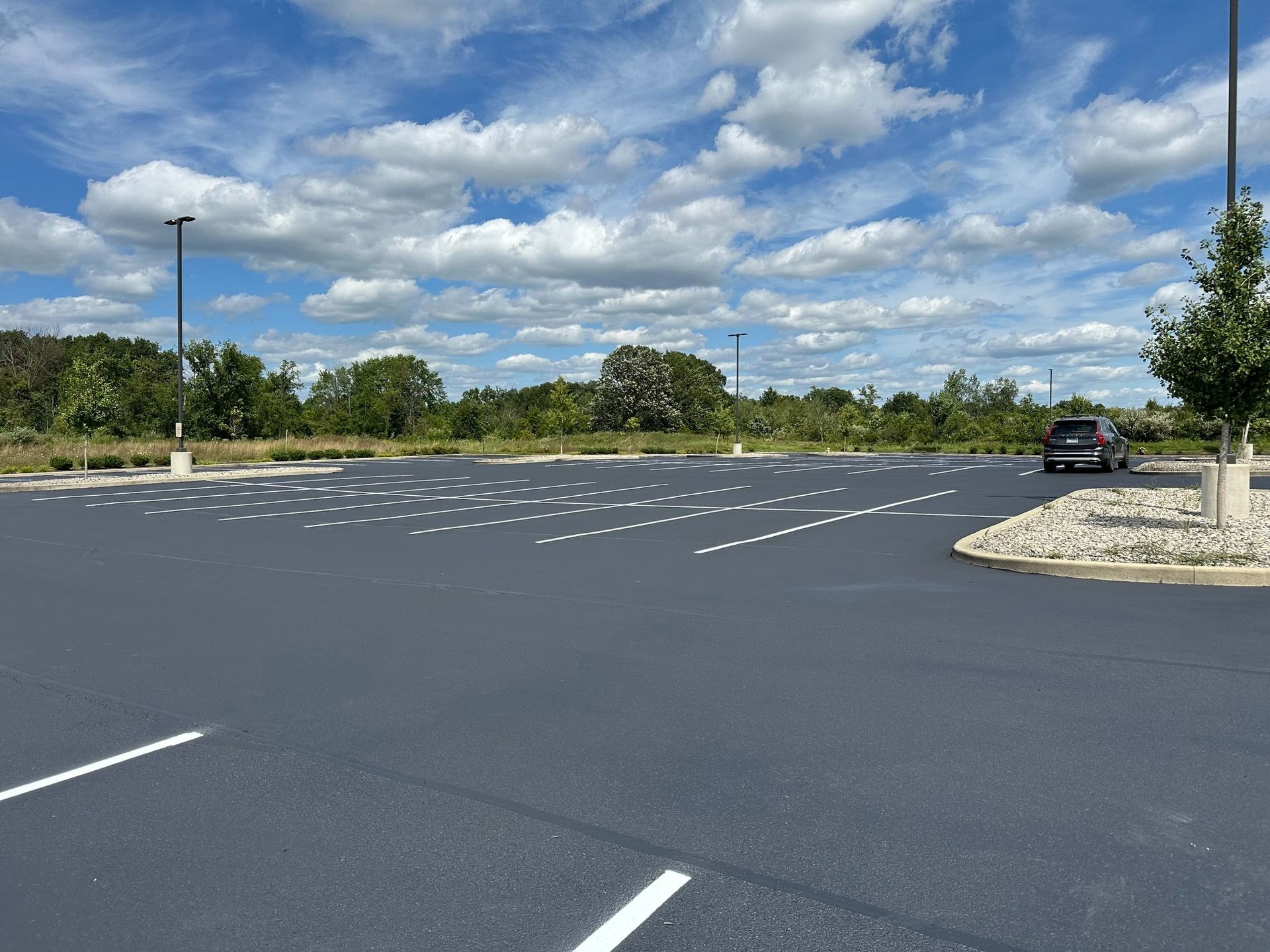 Empty asphalt parking lot with white parking space lines under a cloudy blue sky. A vehicle is parked near trees.