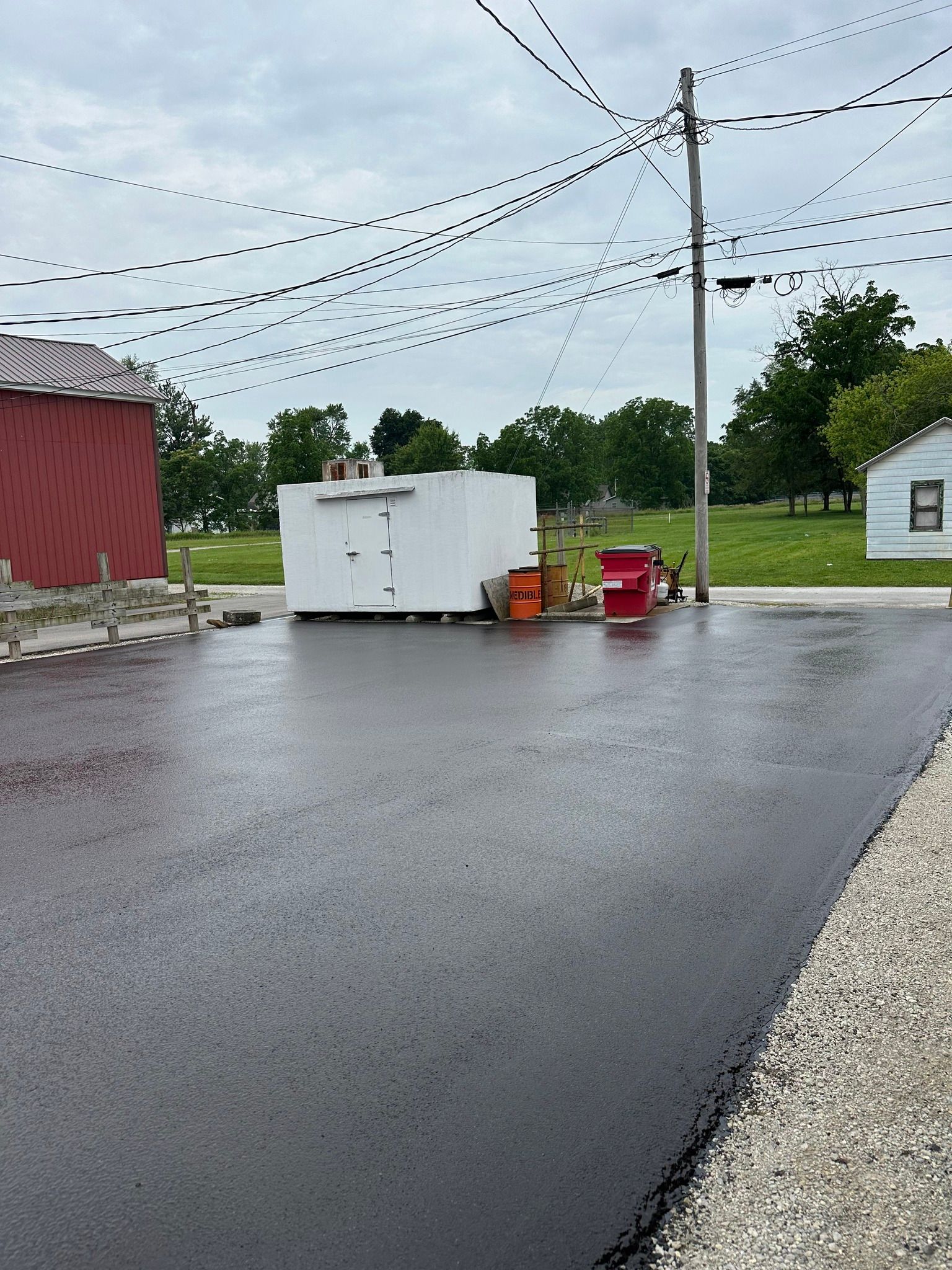 A freshly paved black asphalt lot beside a red barn, with a white storage unit, red bin, and string lights overhead.