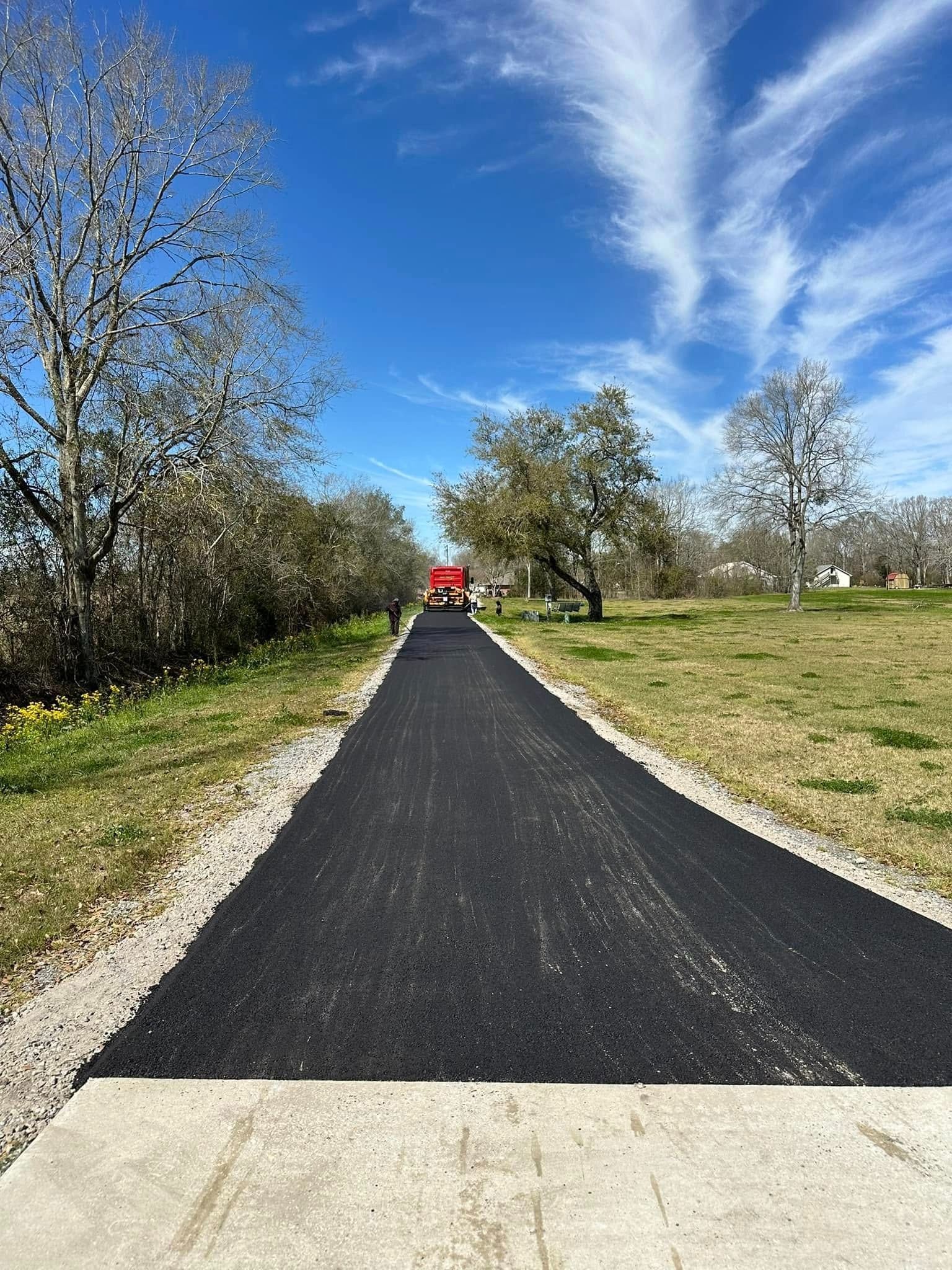 Newly paved black asphalt path with light gray stone edges, green grass, trees, and bright blue sky.
