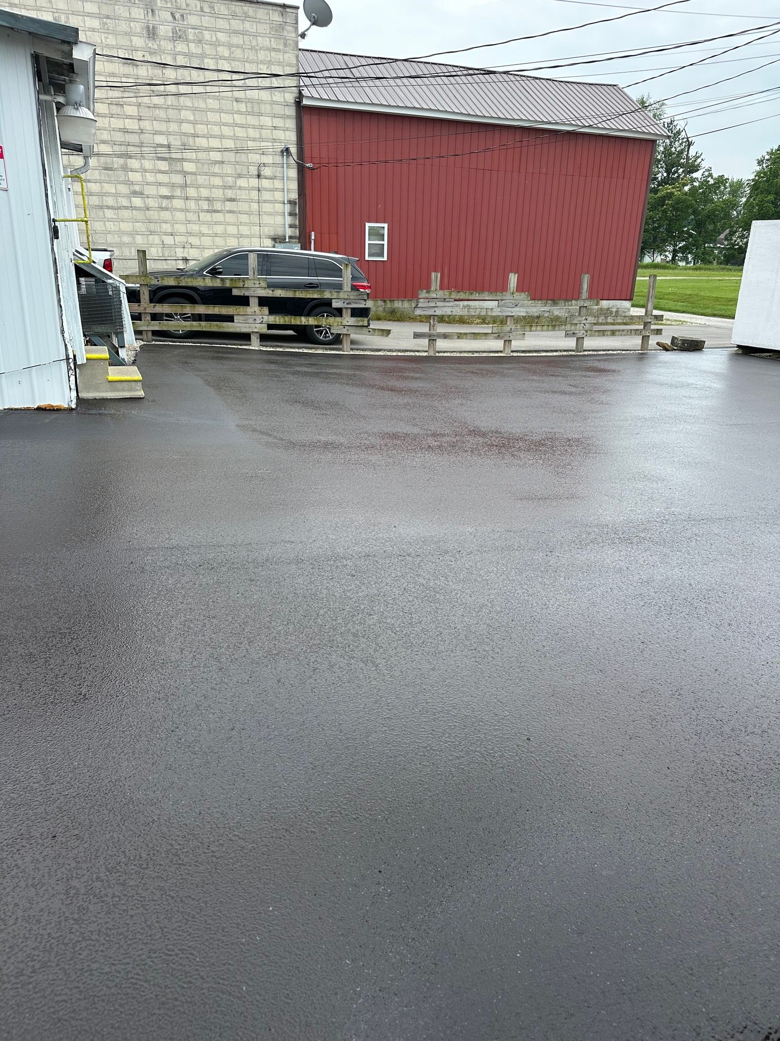 A wet, paved parking area in front of a stone building, a red barn with a white letter E, and a parked black truck.
