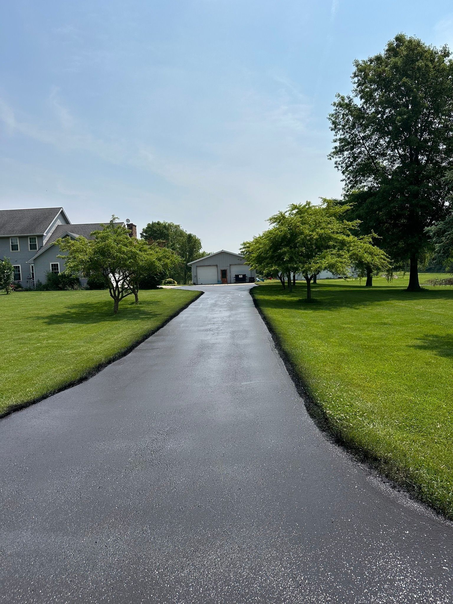 A freshly paved dark asphalt driveway leads through a sunny green lawn towards a house and garage under a clear sky.