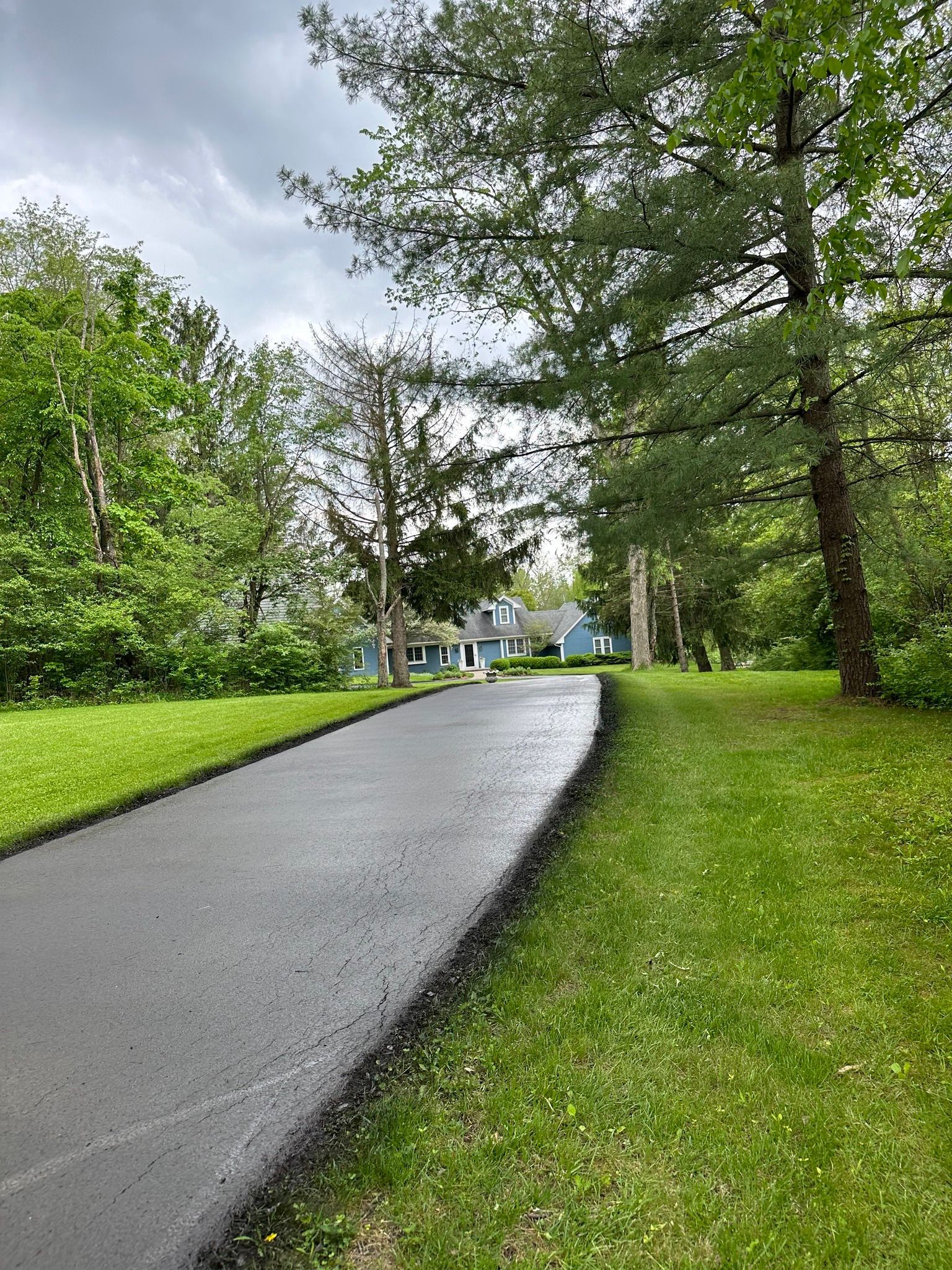 A freshly paved asphalt driveway winds through a grassy residential yard toward a house under a cloudy sky.