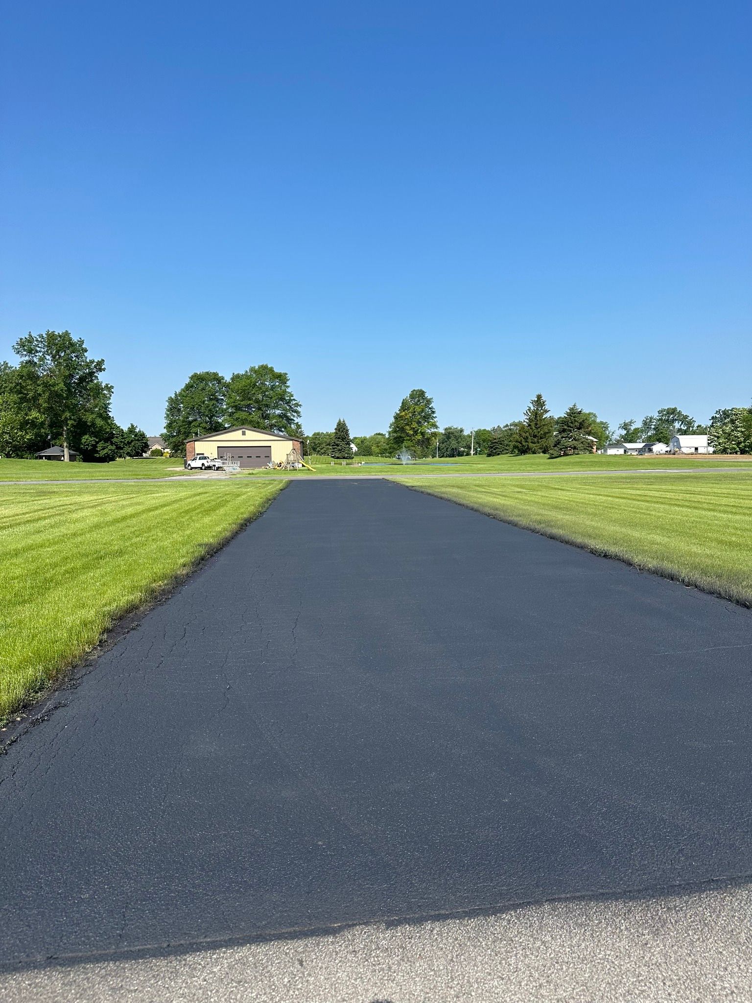 A newly paved asphalt driveway leads through a grassy field toward a garage under a clear blue sky.