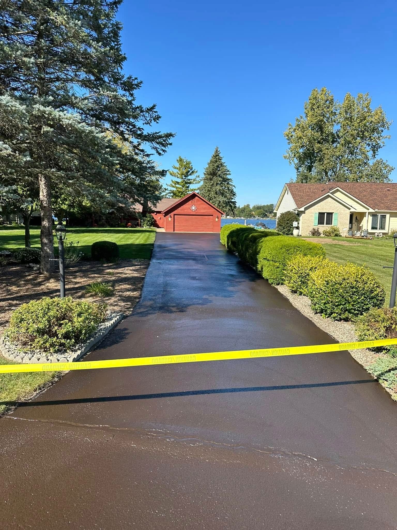 Black asphalt driveway with yellow caution tape, leading to a red shed. Houses and trees flank the driveway under a blue sky.